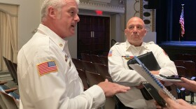 A man with white hair, and wearing a white uniform shirt, speaks to reporters holding notebooks and recorders. Next to him, holding a binder, and listening, is a bald man, wearing the same uniform. Behind him you can see the large auditorium of the Bloomington Center for the Performing Arts.
