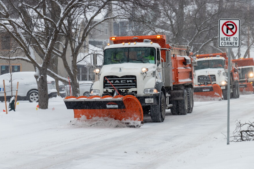 A line of three snow plows on a snow-covered city street.