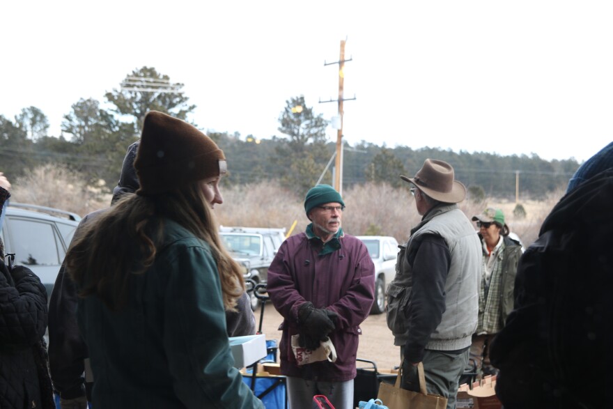 Jo Buckley in a brown beanie and green jacket heads outside to talk with people who are waiting in line at the food pantry. There are two men talking with one another in the line.