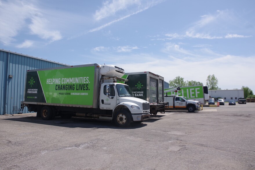 Several trucks with green ang grey designs on them are parked at a loading dock. The truck closest to the camera reads "South Michigan Food Bank, Helping Communities. Changing Lives"