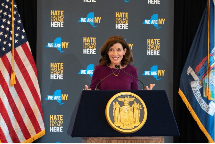 A woman stands at a podium with the American flag on her right and the New York flag on her left.