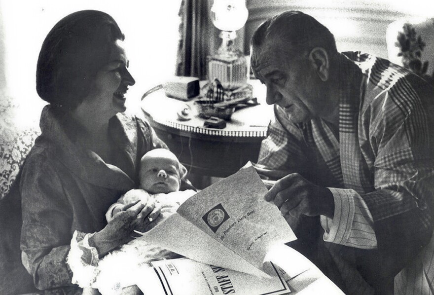 President Lyndon Johnson reads the President's Daily Briefing as his wife Lady Bird Johnson holds their first grandchild in the White House.