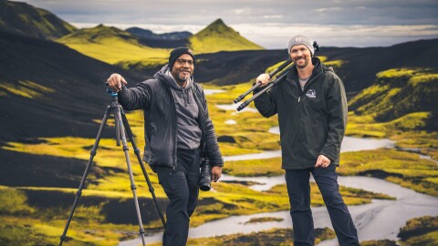 Two men pose with camera gear in a mountain landscape.