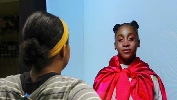 A girl looks at a lifesize holograph of younger Black girl with a cape.