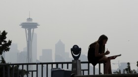 A girl works on a drawing next to an unused viewing scope as a smoky haze obscures the Space Needle and downtown Seattle behind. 