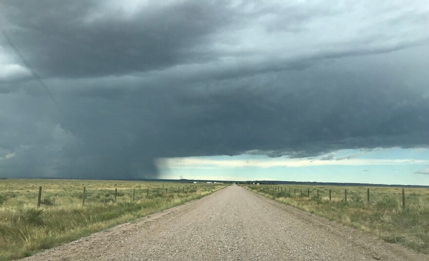 Heavy gray clouds over a dirt road that cuts through a prairie. Rainfall can be seen moving in from the left side.