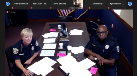 Two police officers in uniform sit on either side of a long, dark table with and pink papers in piles