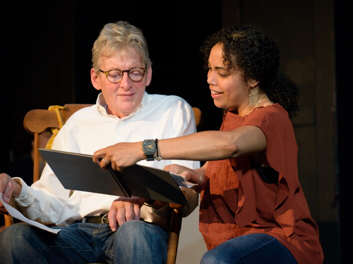 A man sitting in a wooden rocking chair and a woman is crouched next to him showing him a book.