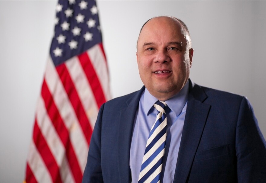 A smiling man with short brown hair, wearing a purple button-down shirt, a blue and white striped tie, and a blue suit jacket. He is standing in front of an American flag.