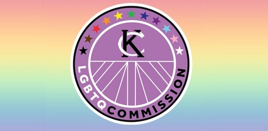 The logo of the L G B T Q Commission of Kansas City, Missouri. A purple circle outlined in white. Arching across the top are 11 stars, each a different color, then a circle outlined in white inside the purple circle containing the linked K and C of Kansas City.  Beneath that inner circle arching along the bottom are the letters L G B T Q in white followed by the word commission in black.