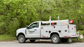 An Ameren Missouri utility truck is parked on a residential city street in Columbia Missouri. Behind the truck the frame is filled with lush green trees. 
