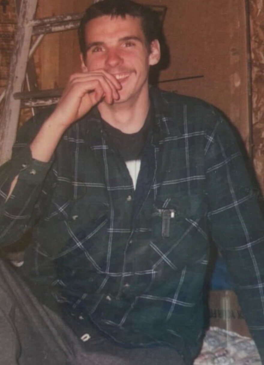 In this old, grainy photo, a young man sits with his leg and head partly out of frame. He's wearing a green shirt and holding a hand up to his mouth, which is smiling.