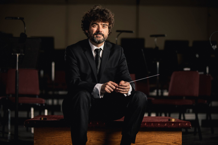 A man in a black suit sitting down, looking forward, holding a conductor's baton