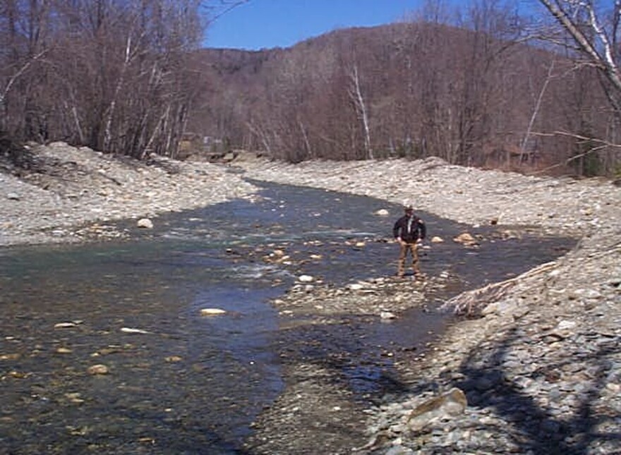 A grainy photo shows a person standing on some rocks surrounded by a large but shallow river.