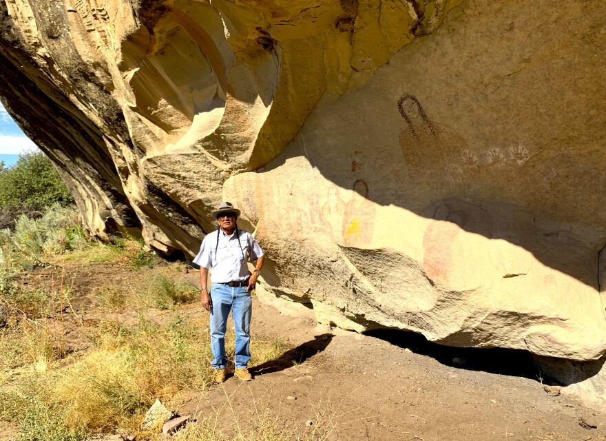 A man stands in front of a rock wall. The rock wall is brown and has drawings of people on it. The man is standing in the sunlight.