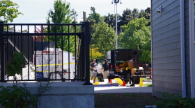 Two individuals can be seen holding a large, plastic object near several trailers in a building parking lot.