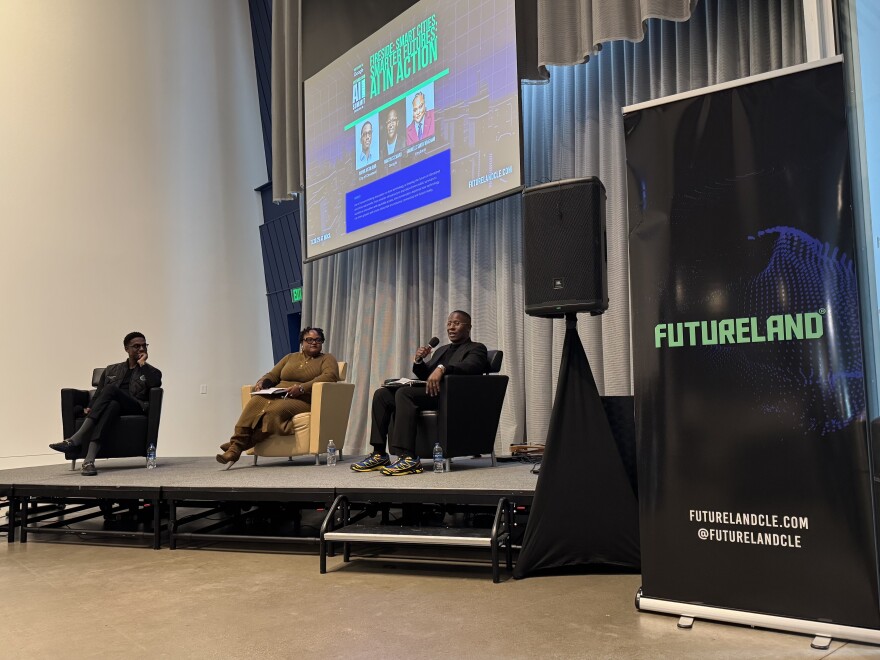 Cleveland Mayor Justin Bibb listens, seated, as Google's Winton Steward speaks on stage at the FutureLAND conference on Artificial Intelligence at Cleveland's Museum of Contemporary Art. Another panelist sits between the two men.