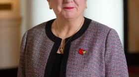 A professional headshot of a woman in a red and black jacket, with a cardinal pin.