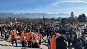 A large crowd of people hold signs in an outdoor space, mountains are visible in the background. Among the crowd is a group of people in shrimp costumes