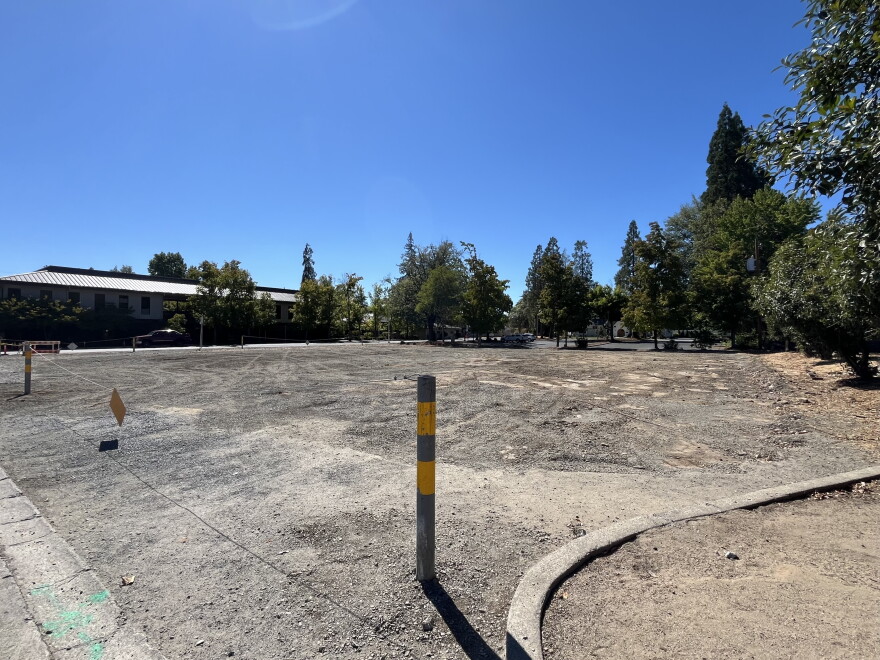 An empty dirt lot is shown, with trees and blue sky in the background. 