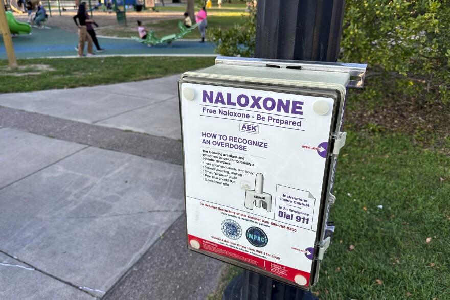 A white, black and red box containing free doses of naloxone, a nasal spray medication to reverse drug overdoses, is installed at on a metal pool in a park. A concrete walking path sits next to it on the left, with childeren at play in the background of the top right. On the other side of the box, a patch of grass, leads to a small bush. 
