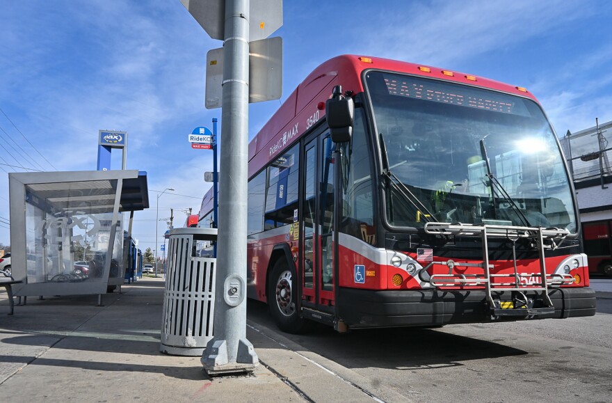 A red commuter bus pulls away from a sidewalk where there is a covered bus stop.