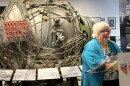 A white-haired, bespectacled woman speaks at a podium. Behind her is a large metal contraption with wires winding around all over. It's a replica of an atom bomb.