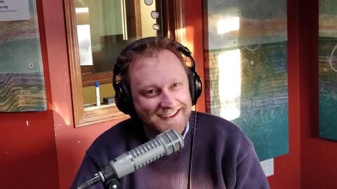 A smiling man wearing headphones sits at a microphone. 