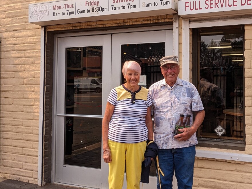 A man and women standing side by side outside the entrance to a store. A sign above the door reads "Taylor's Sausage" with hours posted next to it.