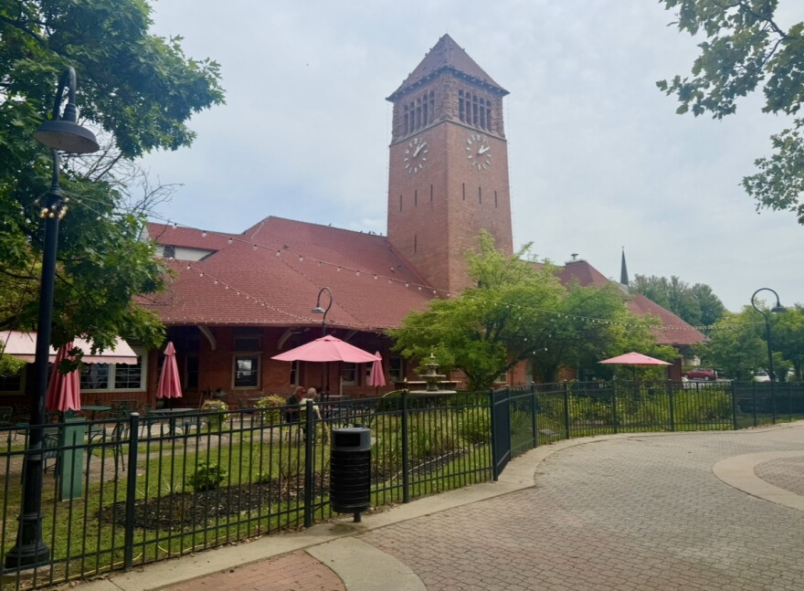 A large red brick building can be seen on a cloudy day. There is a large, fenced-in courtyard in front of he building with a grassy lawn. On the front of the building is a large turret with a clock face on it. 