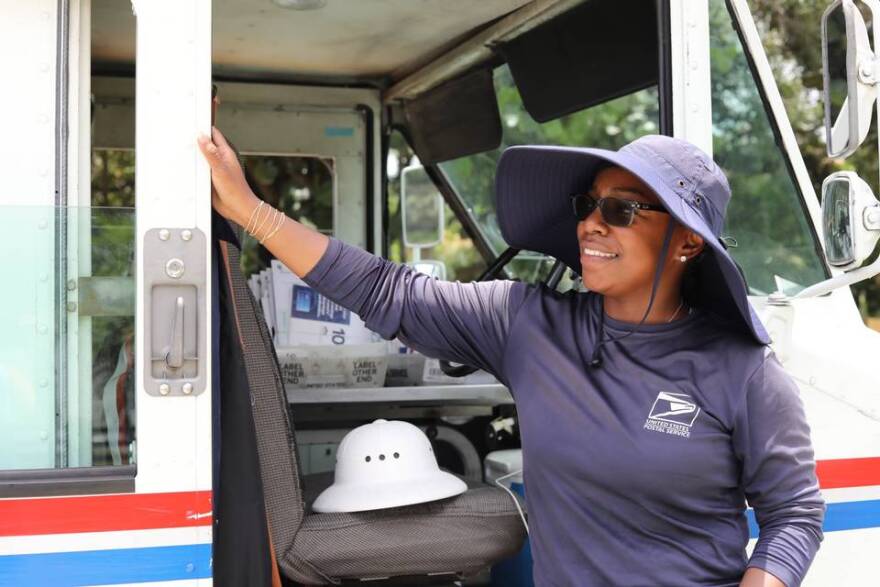 Olivia Gordon, a mail carrier for the United States Postal Service, stands for a portrait alongside her un-air-conditioned mail truck, wearing the floppy hat and long-sleeved shirt that keeps her out of the sun, on July 5, 2023.