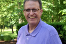  A man wearing glasses smiles at the camera while standing outdoors in front of trees.