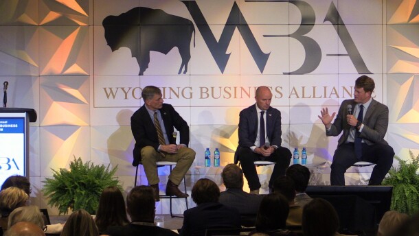 Three men in suits sit in chairs, talking on a stage, with an audience watching.