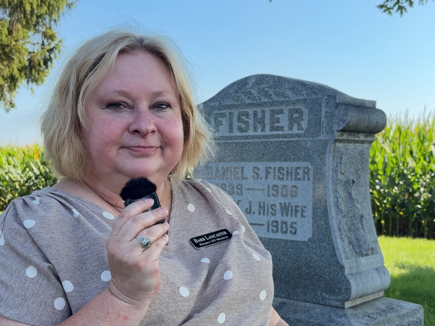 a woman with short blonde hair sits in front of a gravestone