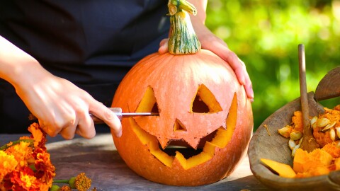 Picture of a Jack-o-lantern that has just been carved .   Facing the camera with a right hand of carver standing behind out of frame is holding knife with blade  finishing the right eye and left hand holding the pumpkin in place at the top. Flowers lower left of image. Wood bowl with pumpkin pulp, partially out of frame lower right of image . 