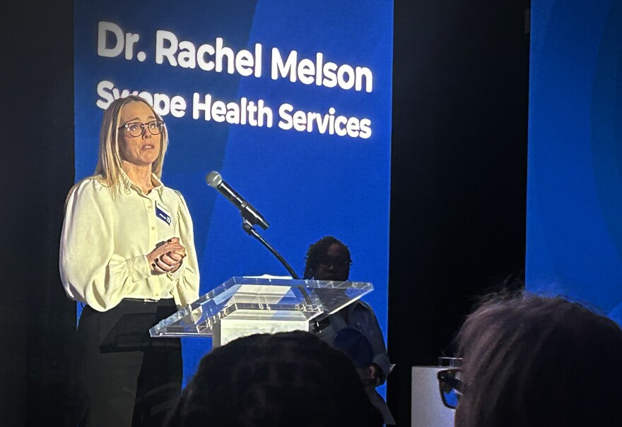 A woman stands at a podium on a stage. Behind her is a blue projection that reads in white letters: "Dr. Rachel Melson, Swope Health Services." The backs of peoples' heads can be seen in the foreground in a darkened auditorium.