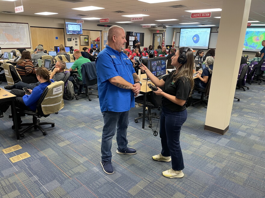 A female reporter interviews a tall white man in a crowded operations center. 