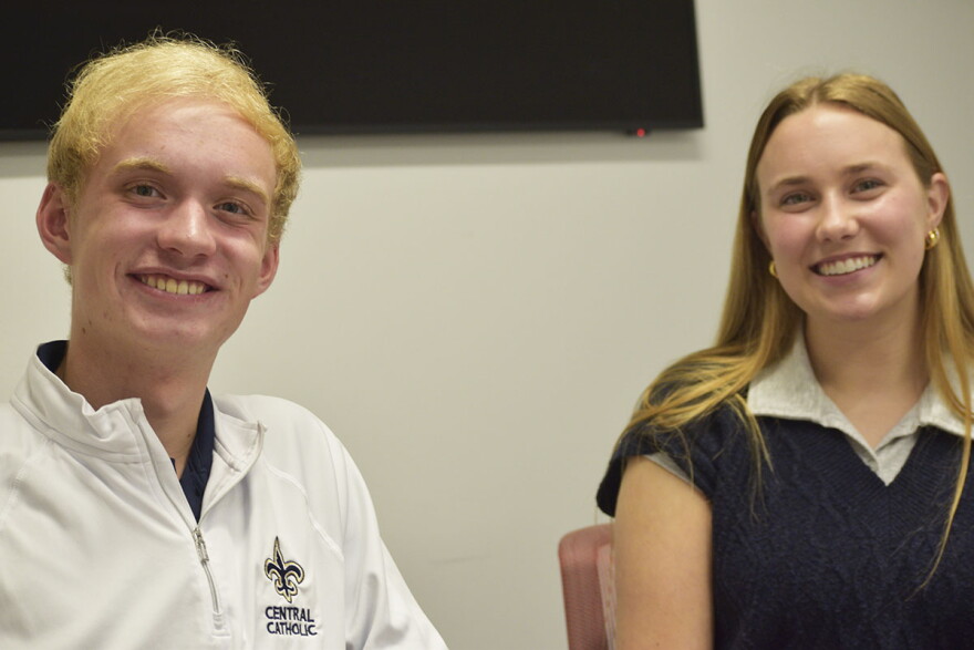 A closeup of a young man in a white quarter-zip jacket with a blue and gold fleur-de-lis logo seated next to a young woman in a blue V-neck sweater and gray t-shirt. Both are smiling and a large television monitor hangs on the wall in the background. 