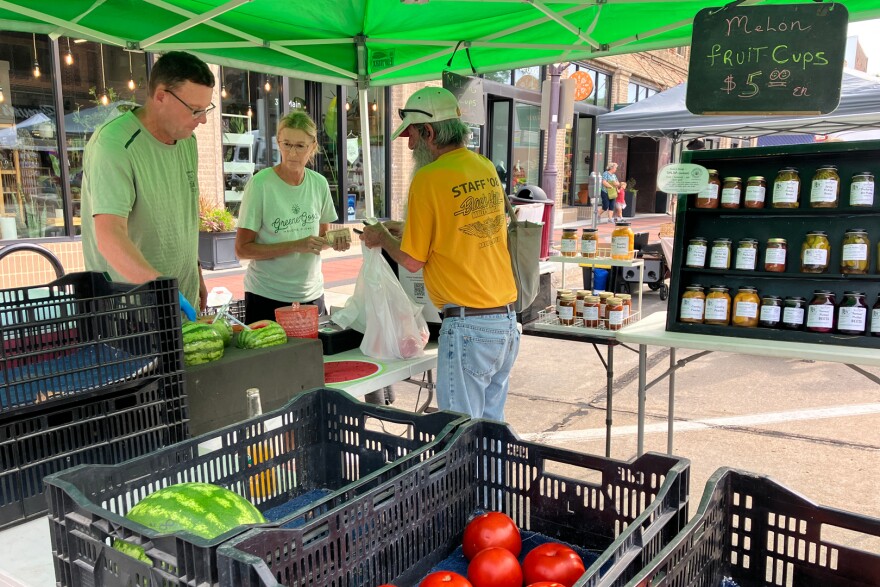 A man in a yellow shirt hands over money to two people selling produce under a tent. A shelf displays pickled beets and other canned produce.