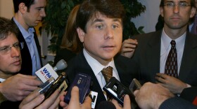 Rod Blagojevich speaks behind several microphones with multiple people crowded around him.