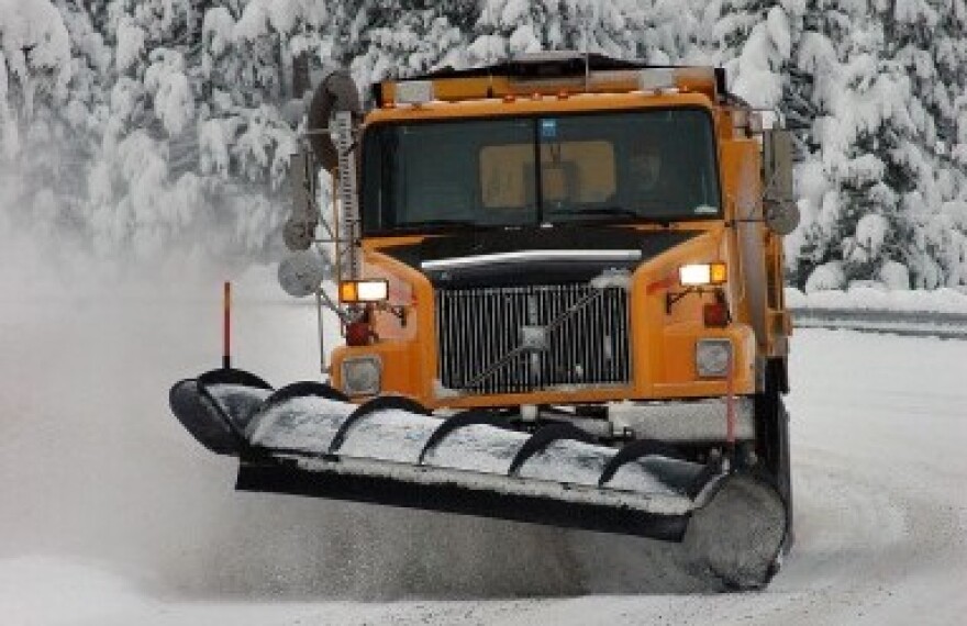 A snow plow clears a road with snow covered tree in the background.