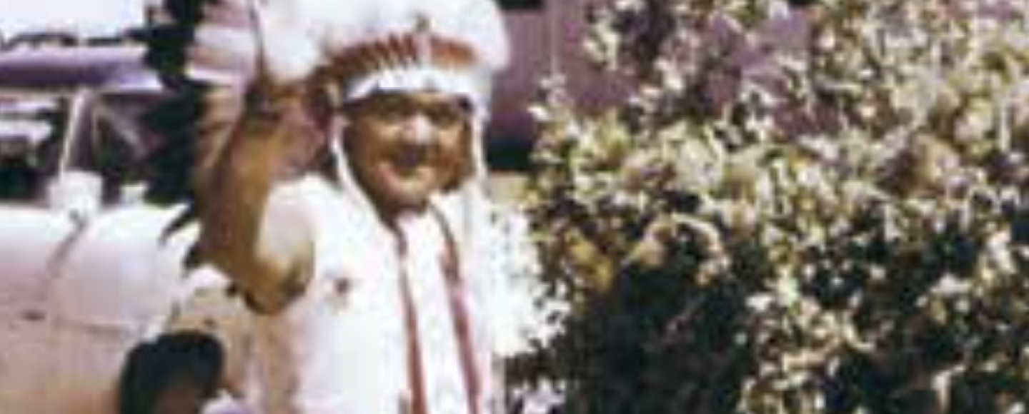 An old photo of a man wearing a headdress waves at the camera.