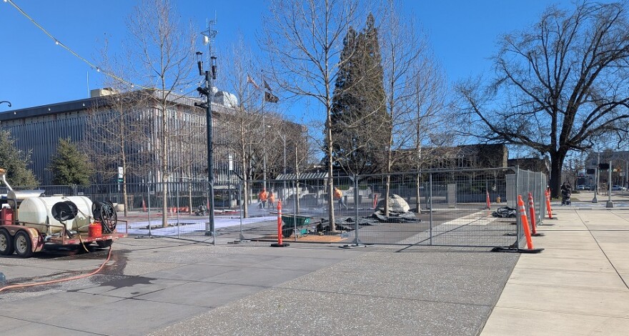 A fenced off area on a concrete plaza. People are working inside the fence. A water truck is nearby.