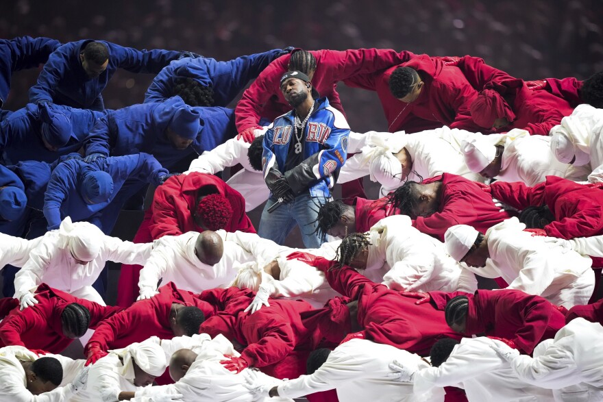 Performer stands on stage, surrounded by dancers in red, white, and blue outfits. The dancers are bent over, forming layered lines. The performer wears a blue and white jersey, jeans, and a black hat. The scene is dramatic and colorful.