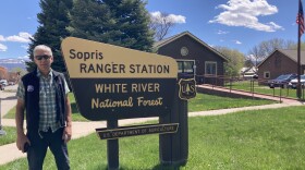 Jim Genung poses outdoors next to a beige and brown sign that reads "Sopris Ranger Station. White River National Forest." 