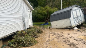 A storage shed that has been knocked over by flood waters sits in a muddy yard.