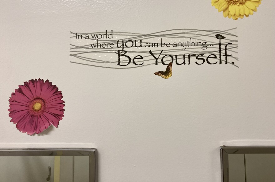 A decal with the stylized words "In a world where you can be anything, Be Yourself," is printed above the mirrors of a bathroom. Two plastic daisies are stuck to the wall next to the quote. 