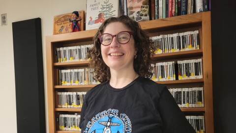 A woman with glasses and curly brown hair smiles in front of a shelf of CDs and wears a black and blue roller derby shirt