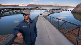 A man wearing a ball cap and jacket stands on a boat ramp in front of a large body of water.