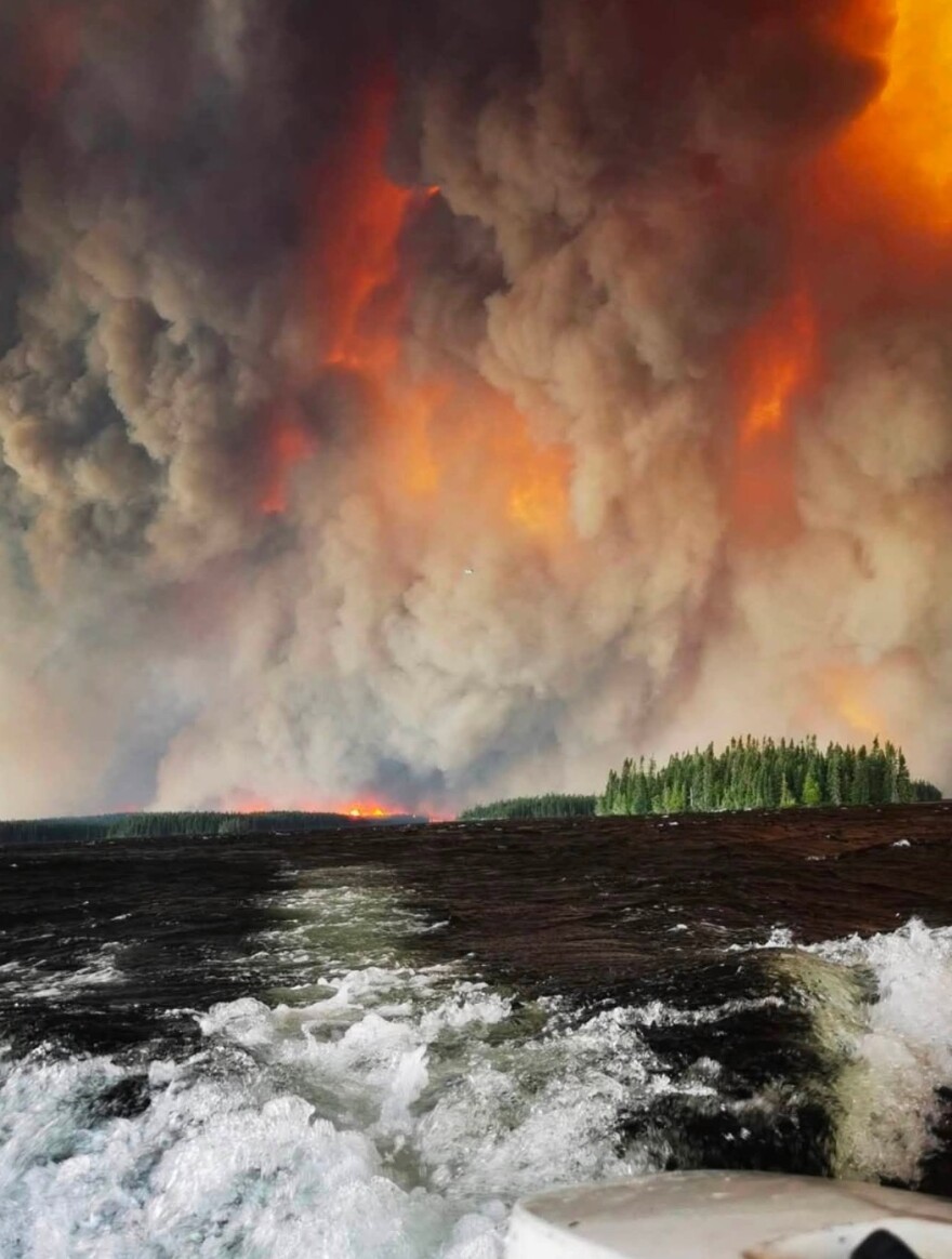 A photo taken from a boat. In the distance, an ominously glowing cloud of gooey black smoke engulfs the entire sky above a forest, looking almost as if the sky was filled with lava from an erupting volcano.